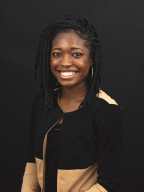 Alice Odama, wearing a khaki and black jacket over a black shirt, poses smiling for a photo in front of a black background.
