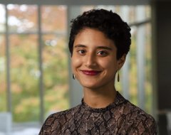 Aya Kamil, wearing a brown and black patterned blouse, smiles for a photo in front of a blurred large window background.