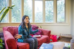 Mehrimo Bakhtalieva, wearing a blue jacket, grey shirt, and grey pants, talks and gestures with her hands while sitting on a red couch in front of a large bay window.