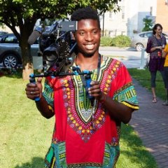 Munya Munyati, wearing a red, yellow, and green shirt while holding a cinema camera on his shoulder, smiles in front of a grassy green background.