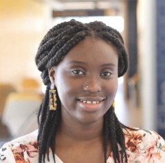 Sandra Ofori, wearing large gold earrings and a red, pink, and white floral shirt, poses smiling for a photo in front of a blurred classroom background.