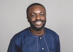 Demi Obayami, wearing a dark blue, collarless, button down, smiles for a headshot in front of a light gray background.