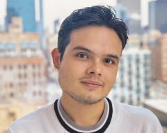 Juan Jose Campos Villalta, wearing a white t-shirt with a black ring collar, poses for a headshot in front of a blurred-out city skyline.