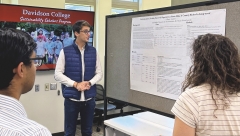 Sandro Chumashvili, wearing a white long-sleeve shirt, black jeans, and a blue puffer vest, stands next to an academic poster mounted on a rolling bulletin board and presents for a small audience.