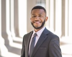 Wilhem Hector, wearing a black suit and tie with a pink button-down, smiles for a headshot in front of a slightly blurred out and shadowed marble background.