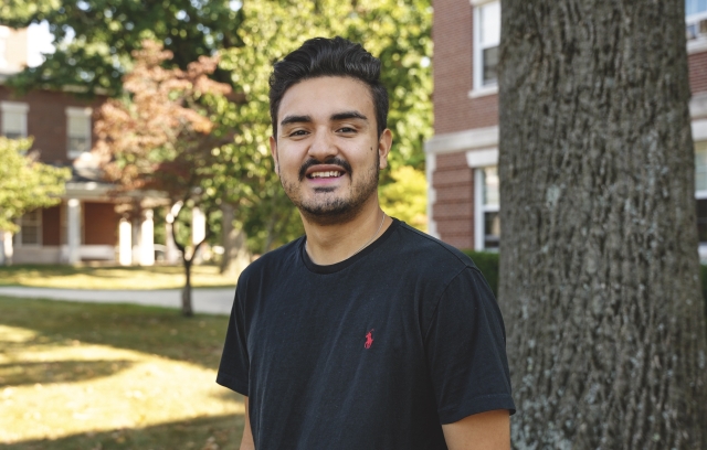 Nico Quijano Franco, wearing a black short-sleeve shirt, smiles for a portrait while standing in front of a tree trunk to his left, and a green lawn leading back to a brick building on his right.