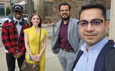 Four college students of various heights, skin tones, and clothing colors pose together for a selfie outside on a street corner.