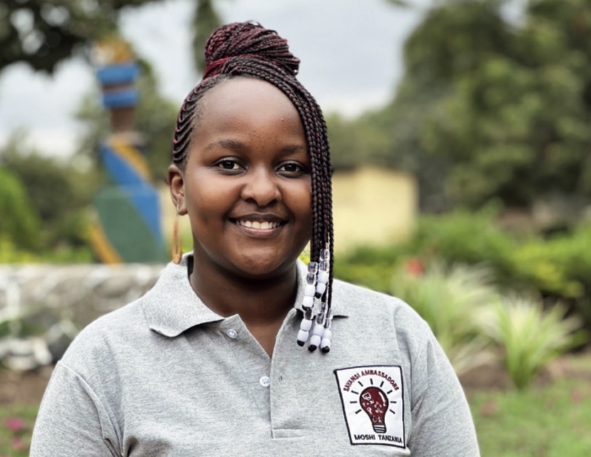 Eli Swai, wearing a gray polo with a breast-pocket patch featuring a red and yellow light bulb, smiles for a headshot in front of a green and lush outdoor garden.