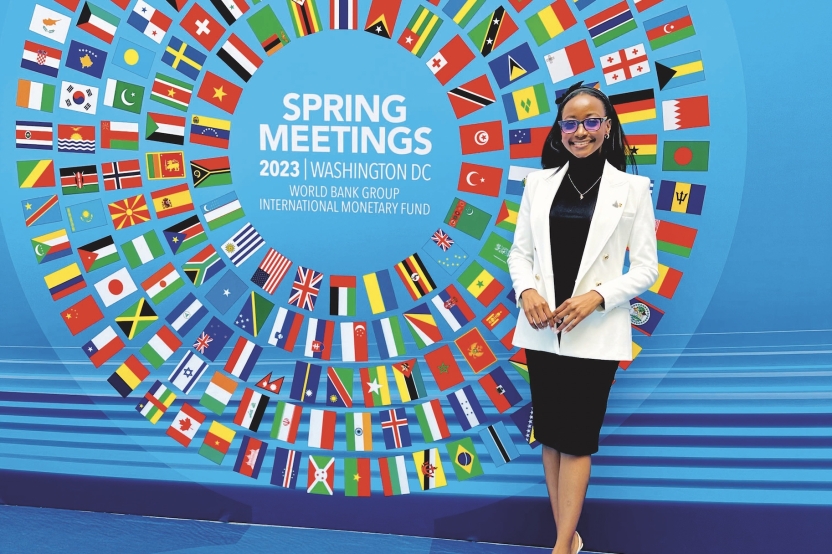 Tatenda Dzvimbo, wearing a white and black pantsuit, poses for a photo next to a mural featuring a swirling circle of global flags on a blue background, and the text "Spring Meetings 2023 Washington D.C."