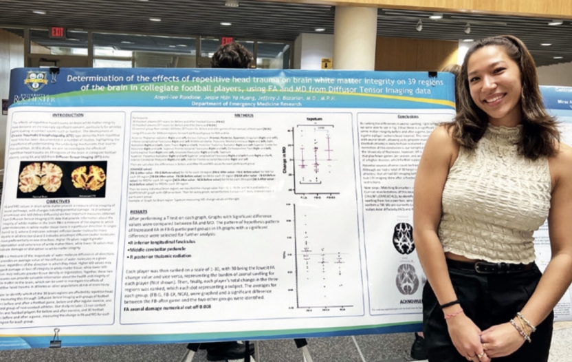 Angel-lee Randone, wearing black pants and a beige tank top, poses to the right of a large academic poster full of charts and pictures of the brain.