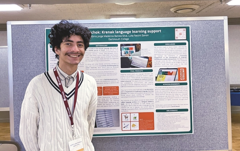 Antonio Jorge Medeiros Batista Silva, wearing a white sweater over a white button-down, smiles posing for a photo in front of a colorful academic poster pinned up to a bulletin board.