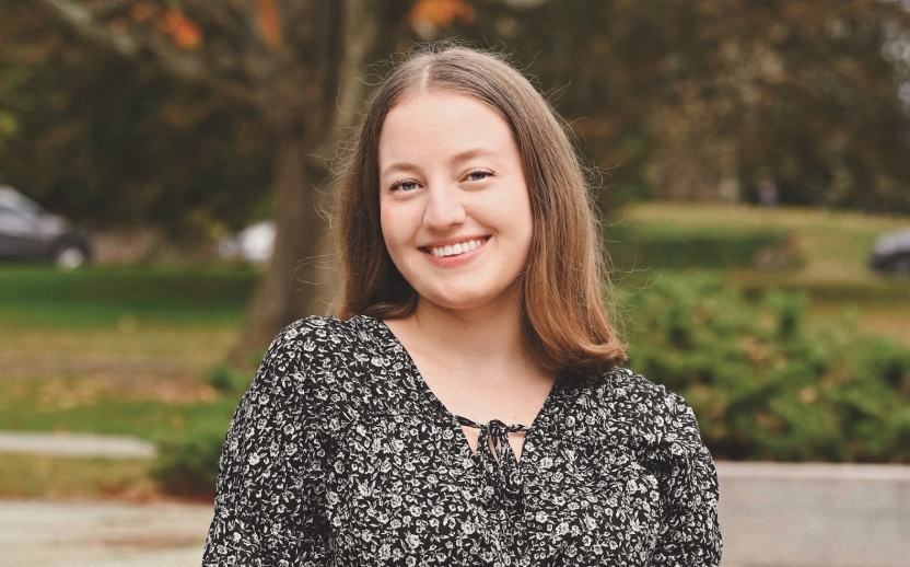 Stella König, wearing a black and white dotted blouse, smiles for a headshot in front of a green lawn and a tree with orange leaves.